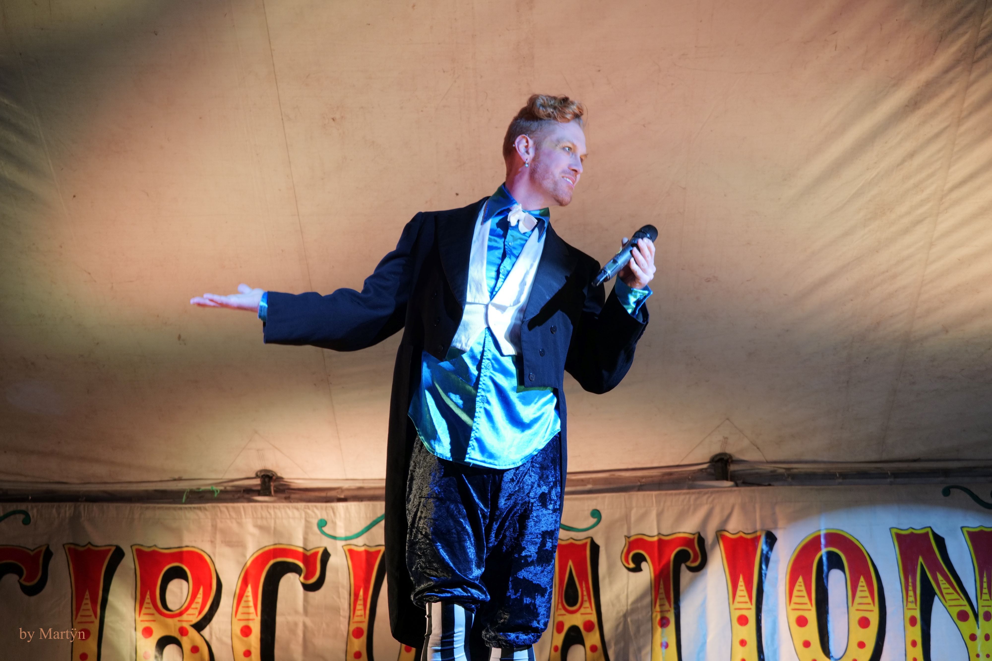 A person in a short tailed circus-ringleader jacket and spiffy clothes, gesturing with one hand with a mic in the other